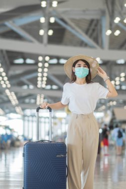 Havaalanında yürüyen yüz maskesi takan genç bir kadın, Coronavirus hastalığı enfeksiyonunu koruyan, şapkalı Asyalı bir kadın gezgin. Aşı artırıcı doz konseptinden sonra seyahat zamanı