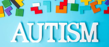 Renkli ahşap yapboz parçalarıyla AUTISM metni, mavi arkaplanda geometrik şekil bloğu. Sağlık, Otistik Spektrum Bozukluğu ve Dünya Otizm Farkındalığı Günü