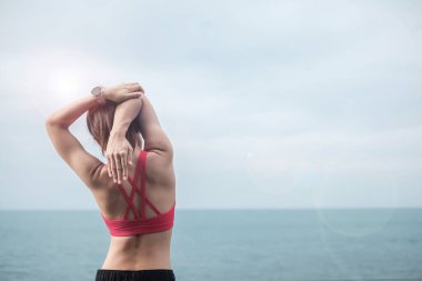 Spor kıyafetli genç bir kadın, vücudu okyanus manzarasına karşı esnetiyor, sabahları sağlıklı kadın egzersizi yapıyor. Spor, sağlık ve iş hayatı dengesi kavramları