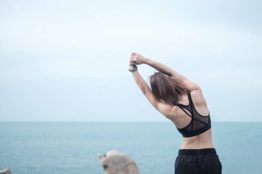 Spor kıyafetli genç bir kadın, vücudu okyanus manzarasına karşı esnetiyor, sabahları sağlıklı kadın egzersizi yapıyor. Spor, sağlık ve iş hayatı dengesi kavramları