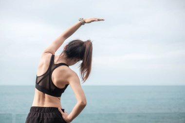 Spor kıyafetli genç bir kadın, vücudu okyanus manzarasına karşı esnetiyor, sabahları sağlıklı kadın egzersizi yapıyor. Spor, sağlık ve iş hayatı dengesi kavramları