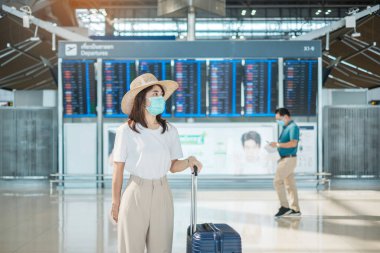 Havaalanında yürüyen yüz maskesi takan genç bir kadın, Coronavirus hastalığı enfeksiyonunu koruyan, şapkalı Asyalı bir kadın gezgin. Yolculuk, Test ve Git ve Aşı Artırıcı Doz Konsepti