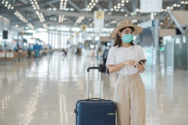 Yüz maskesi takan ve havaalanında cep telefonu kullanan Asyalı kadın turist Coronavirus hastalığı enfeksiyonunu koruyor. Aşı artırıcı doz konseptinden sonra seyahat zamanı