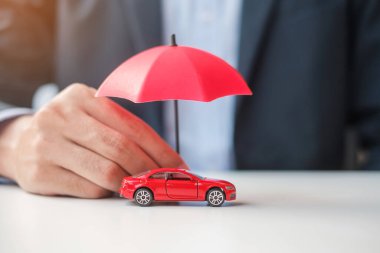 İş adamı şemsiyesini tutuyor ve masada kırmızı araba oyuncağını örtüyor. Araba sigortası, garanti, onarım, finans, bankacılık ve para kavramı