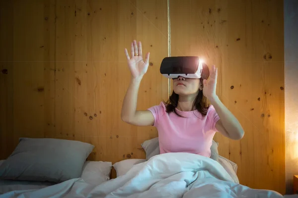 Young woman using virtual reality headset in bed. VR, Future digital technology, game, movies entertainment, metaverse, NFT and 3D cyberspace concept