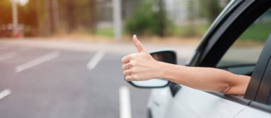 Kadın, yolda araba kullanırken başparmağını kaldırıyor, modern elektrikli otomobilde direksiyonu elle kontrol ediyor. Yolculuk, yolculuk ve güvenlik ulaşım kavramları