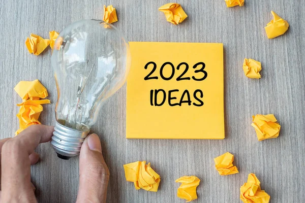 2023 Idea words on yellow note and crumbled paper with Businessman holding lightbulb on wooden table background. New Year New Start Creative, Innovation, Imagination, Resolution and Goal concept