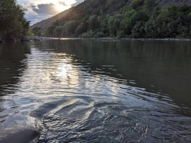 Gün batımında nehrin üzerinde güzel bulutlu bir gökyüzü. Nehrin yüksek kıyısı otlarla kaplıdır. Nehrin karşı kıyısı alçak ve üzerinde bir orman yetişiyor..