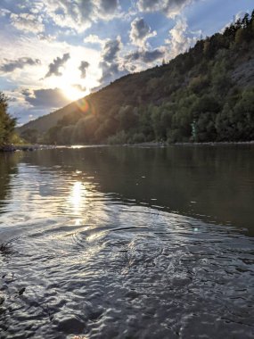 Gün batımında nehrin üzerinde güzel bulutlu bir gökyüzü. Nehrin yüksek kıyısı otlarla kaplıdır. Nehrin karşı kıyısı alçak ve üzerinde bir orman yetişiyor..