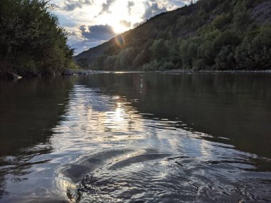 Gün batımında nehrin üzerinde güzel bulutlu bir gökyüzü. Nehrin yüksek kıyısı otlarla kaplıdır. Nehrin karşı kıyısı alçak ve üzerinde bir orman yetişiyor..