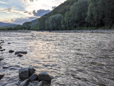 Gün batımında nehrin üzerinde güzel bulutlu bir gökyüzü. Nehrin yüksek kıyısı otlarla kaplıdır. Nehrin karşı kıyısı alçak ve üzerinde bir orman yetişiyor..