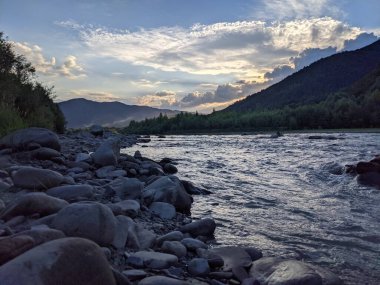 Gün batımında nehrin üzerinde güzel bulutlu bir gökyüzü. Nehrin yüksek kıyısı otlarla kaplıdır. Nehrin karşı kıyısı alçak ve üzerinde bir orman yetişiyor..
