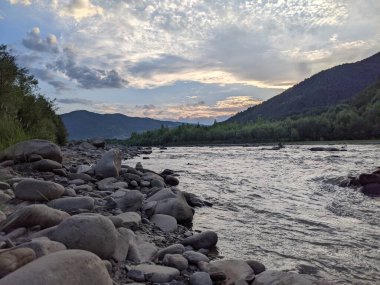 Gün batımında nehrin üzerinde güzel bulutlu bir gökyüzü. Nehrin yüksek kıyısı otlarla kaplıdır. Nehrin karşı kıyısı alçak ve üzerinde bir orman yetişiyor..