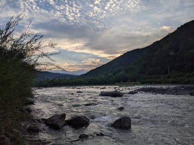 Gün batımında nehrin üzerinde güzel bulutlu bir gökyüzü. Nehrin yüksek kıyısı otlarla kaplıdır. Nehrin karşı kıyısı alçak ve üzerinde bir orman yetişiyor..