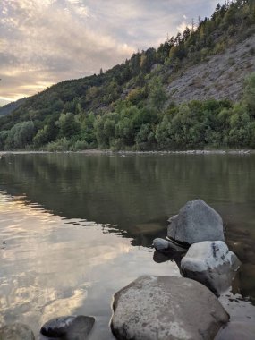 Gün batımında nehrin üzerinde güzel bulutlu bir gökyüzü. Nehrin yüksek kıyısı otlarla kaplıdır. Nehrin karşı kıyısı alçak ve üzerinde bir orman yetişiyor..