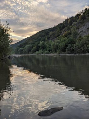 Gün batımında nehrin üzerinde güzel bulutlu bir gökyüzü. Nehrin yüksek kıyısı otlarla kaplıdır. Nehrin karşı kıyısı alçak ve üzerinde bir orman yetişiyor..