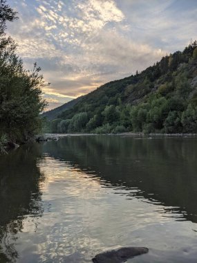Gün batımında nehrin üzerinde güzel bulutlu bir gökyüzü. Nehrin yüksek kıyısı otlarla kaplıdır. Nehrin karşı kıyısı alçak ve üzerinde bir orman yetişiyor..