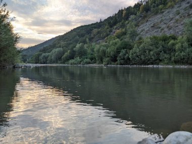 Gün batımında nehrin üzerinde güzel bulutlu bir gökyüzü. Nehrin yüksek kıyısı otlarla kaplıdır. Nehrin karşı kıyısı alçak ve üzerinde bir orman yetişiyor..