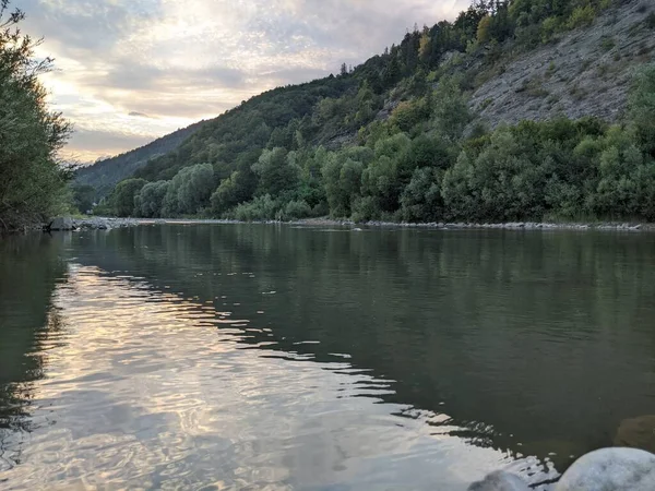 Gün batımında nehrin üzerinde güzel bulutlu bir gökyüzü. Nehrin yüksek kıyısı otlarla kaplıdır. Nehrin karşı kıyısı alçak ve üzerinde bir orman yetişiyor..