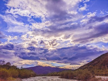 Gün batımında nehrin üzerinde güzel bulutlu bir gökyüzü. Nehrin yüksek kıyısı otlarla kaplıdır. Nehrin karşı kıyısı alçak ve üzerinde bir orman yetişiyor..
