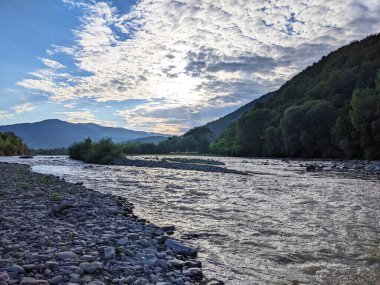 Gün batımında nehrin üzerinde güzel bulutlu bir gökyüzü. Nehrin yüksek kıyısı otlarla kaplıdır. Nehrin karşı kıyısı alçak ve üzerinde bir orman yetişiyor..