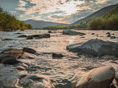 Gün batımında nehrin üzerinde güzel bulutlu bir gökyüzü. Nehrin yüksek kıyısı otlarla kaplıdır. Nehrin karşı kıyısı alçak ve üzerinde bir orman yetişiyor..