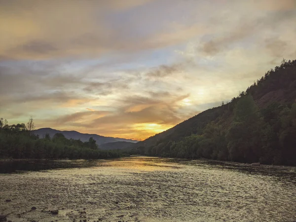 Beautiful cloudy sky at sunset over the river. The high bank of the river is overgrown with grass. The opposite bank of the river is low and there is a forest growing on it.