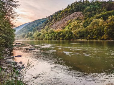 Gün batımında nehrin üzerinde güzel bulutlu bir gökyüzü. Nehrin yüksek kıyısı otlarla kaplıdır. Nehrin karşı kıyısı alçak ve üzerinde bir orman yetişiyor..
