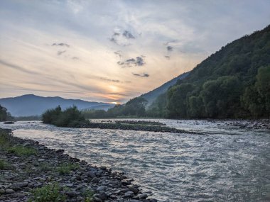 Gün batımında nehrin üzerinde güzel bulutlu bir gökyüzü. Nehrin yüksek kıyısı otlarla kaplıdır. Nehrin karşı kıyısı alçak ve üzerinde bir orman yetişiyor..