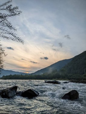 Gün batımında nehrin üzerinde güzel bulutlu bir gökyüzü. Nehrin yüksek kıyısı otlarla kaplıdır. Nehrin karşı kıyısı alçak ve üzerinde bir orman yetişiyor..