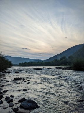 Gün batımında nehrin üzerinde güzel bulutlu bir gökyüzü. Nehrin yüksek kıyısı otlarla kaplıdır. Nehrin karşı kıyısı alçak ve üzerinde bir orman yetişiyor..