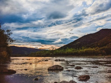 Gün batımında nehrin üzerinde güzel bulutlu bir gökyüzü. Nehrin yüksek kıyısı otlarla kaplıdır. Nehrin karşı kıyısı alçak ve üzerinde bir orman yetişiyor..