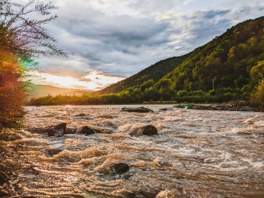 Beautiful cloudy sky at sunset over the river. The high bank of the river is overgrown with grass. The opposite bank of the river is low and there is a forest growing on it.