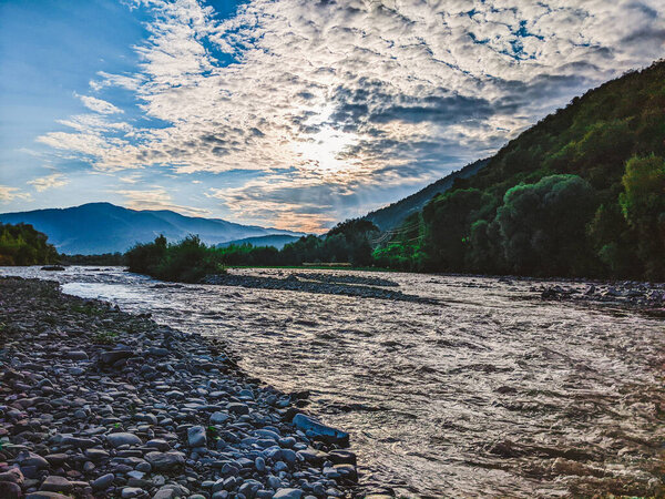 Beautiful cloudy sky at sunset over the river. The high bank of the river is overgrown with grass. The opposite bank of the river is low and there is a forest growing on it.