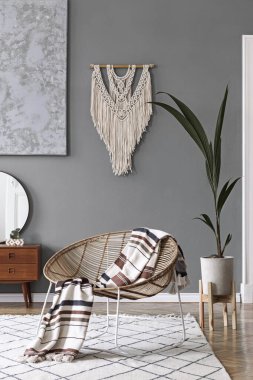 Kahve masası, rattan koltuğu, bitkiler, halı ve güzel boho aksesuarları olan yaratıcı ve rahat bir oturma odası kompozisyonu. Gri duvarlar ve parke zemin.