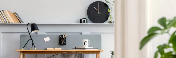Scandinavian home office interior with wooden desk, design chair, wood panleing with shelf, plant, table lamp, office supplies and elegant accessories in modern home decor.