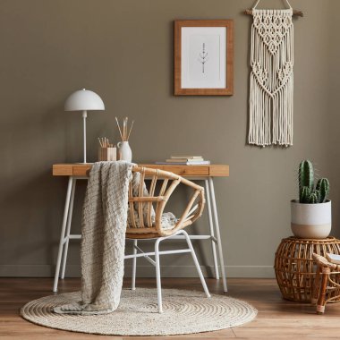 Ev ofis odasının içinde modern kompozisyon ahşap masa, şık koltuk, bambu raf, halı, makrome, poster çerçevesi, ofis malzemeleri, dekorasyon ve kişisel aksesuarlar..