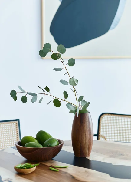 Minimalist composition on the design wooden table with fruits, tropical leaf in vase, abstract painitngs and stylish chair. Modern dining room. Template.