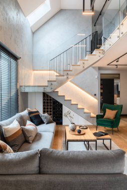Oturma odasının içindeki merdivenlerin şık kompozisyonu. Gri kanepe, yeşil kadife koltuk, sehpa ve minimalist kişisel aksesuarlar. Modern ev dekorasyonu. Şablon.