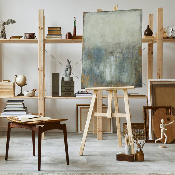 Unique artist workspace interior with stylish teak commode, wooden easel, bookcase, artworks, painting accessories, decoration and elegant personal stuff. Modern work room for artist. Template. 