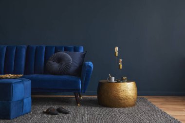 Görkemli mavi kadife kanepe, puf, altın metal yan masa ve modern ev aksesuarları ile modern oturma odası iç tasarımı. Koyu mavi duvar. Şablon. Boşluğu kopyala.