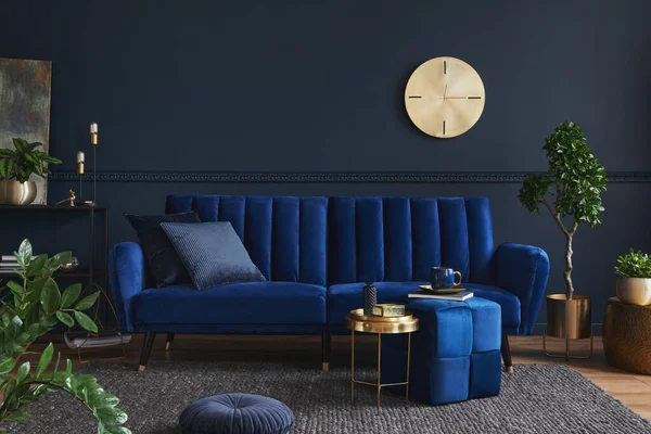 Creative compositon of modern living room interior design with glamour blue sofa, metal shelf, coffee table and elegant home accessories. Dark blue wall. Home staging. Template. Copy space