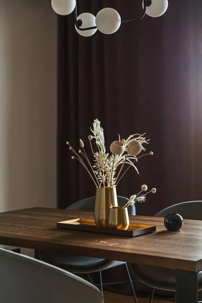 Modern and vintage dining room interior with brown wooden table, grey chairs and stylish chandelier. White wall and brown curtains. Boho and minimalistic accessories. Gold metalic vases in metalic tray. Dried flowers.