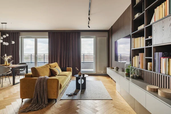 Creative and modern vintage living room interior design with yellow sofa and coffee table on grey carpet. Lamella wall with tv, bookcase and chest of drawers . Herringbone parquet., Brown curtain in windows. Template.