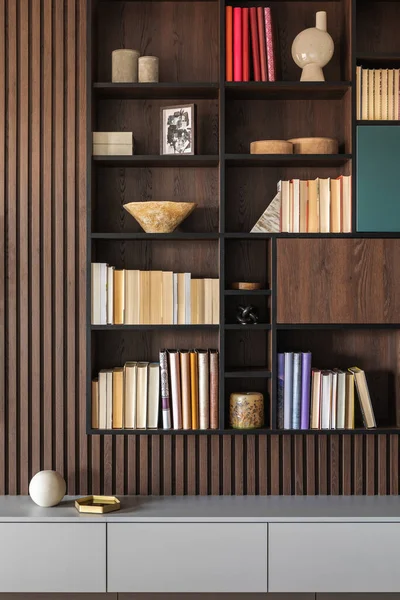 Modern bookcase in lamells wall. Grey chest of drawers. Bookcase a lot of books and accessories. Herringbone wooden parquet. Cat in open spaces.