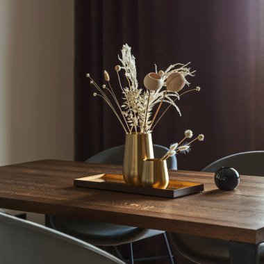 Modern ve klasik yemek odası. Kahverengi ahşap masa, gri sandalyeler ve şık avizeler. Beyaz duvar ve kahverengi perdeler. Boho ve minimalist aksesuarlar. Metal tepside altın metalik vazolar. Kurumuş çiçekler. 