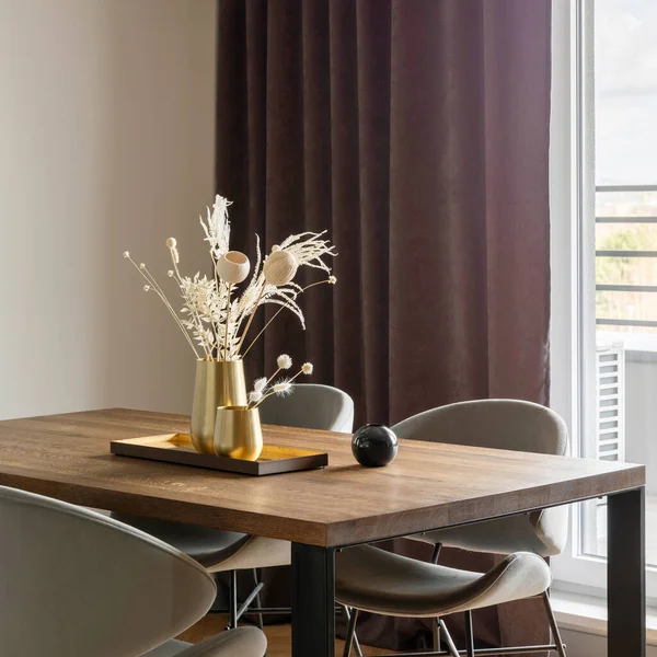 Modern and vintage dining room interior with brown wooden table, grey chairs and stylish chandelier. White wall and brown curtains. Boho and minimalistic accessories. Gold metalic vases in metalic tray. Dried flowers.