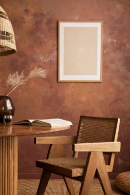 Stylish boho vintage dining room interior with poster mock up. Round table with rattan armchair, vase with dried flowers and cup. Brown wall and rattan lamp. Mock up poster. Template. 