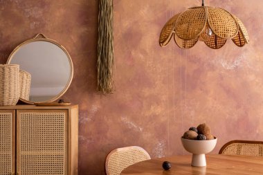 Stylish boho vintage dining room interior with round table, rattan chair, commode and accessories. Brown wall and rattan lamp. Template. 