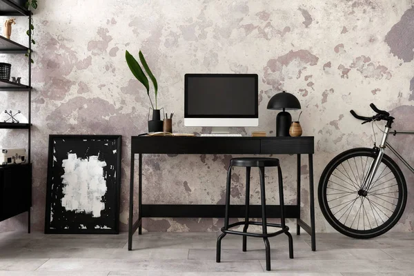 Concrete interior of home office with black desk, brown armchair, computer screen, office accessories,lamp and bicycle. Rack with personal accessories. Home decor. Template.